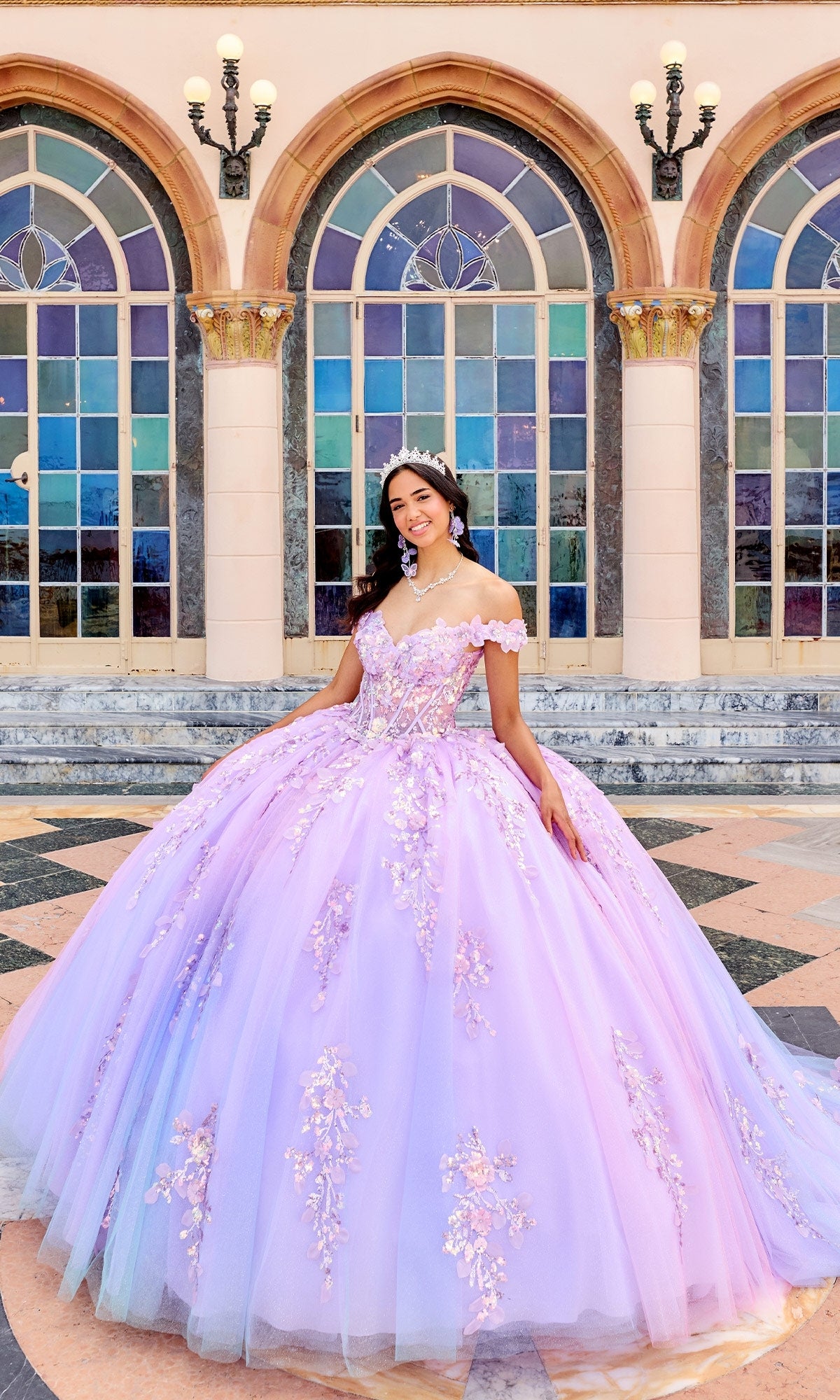A young woman smiles in front of arched windows and columns, wearing the Quinceanera Dress PR30162 by Princesa—a lavender ball gown with floral details and an off-the-shoulder neckline.